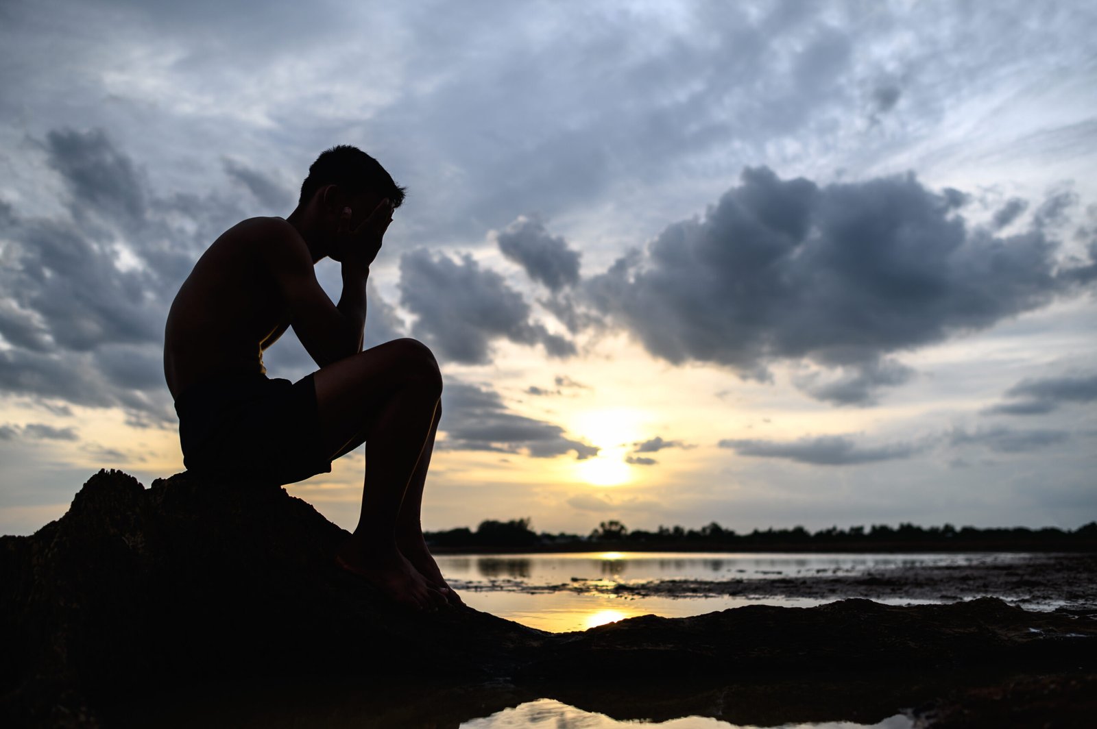 A man sat bent his knees, holding his hands on the face on the base of the tree and there is water around.